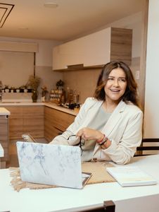A woman in a cozy kitchen setting, smiling while working on a laptop and holding glasses.