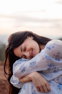 A woman in a floral dress smiling peacefully outdoors at sunset, embracing tranquility.