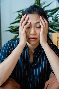 A woman sitting indoors, holding her head in stress or worry, surrounded by plants.